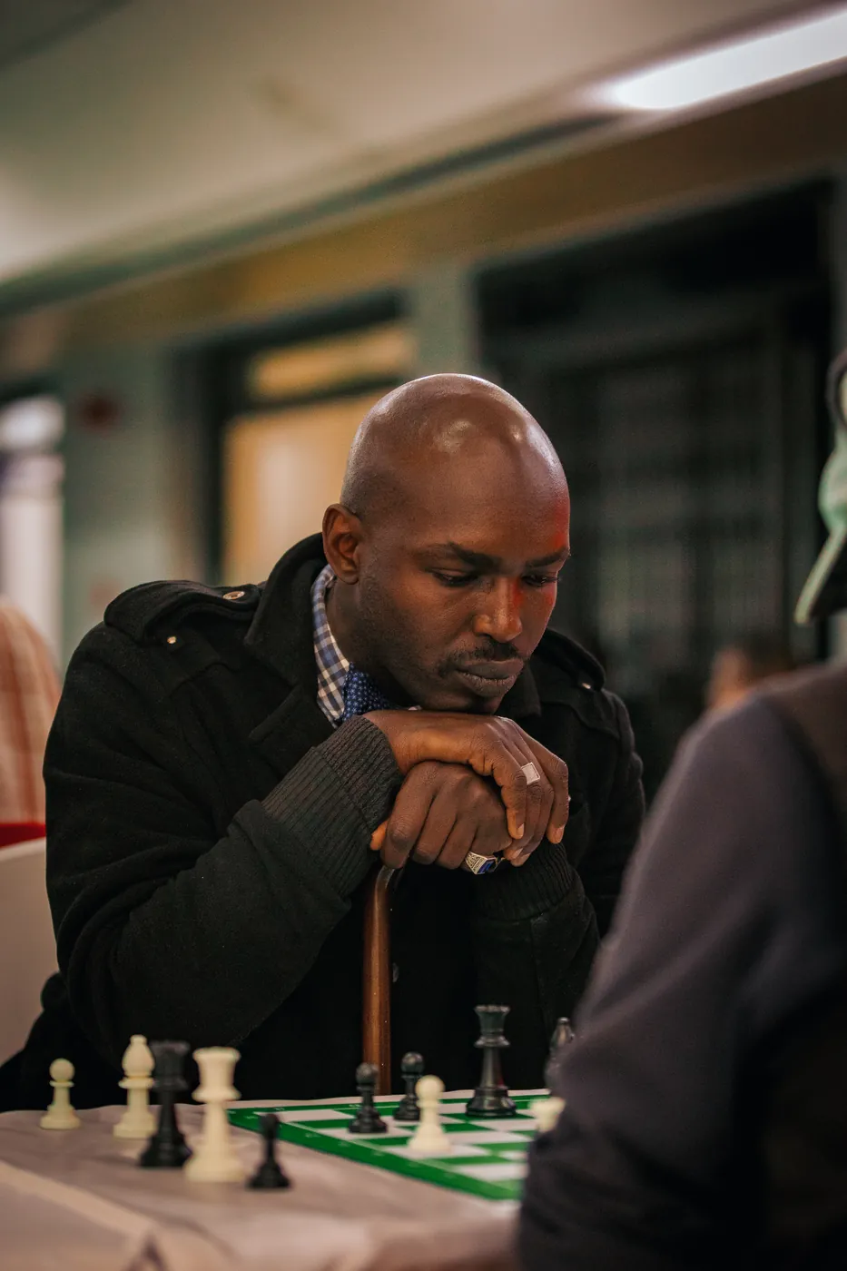 A bald player in a dark coat rests on his cane over the board