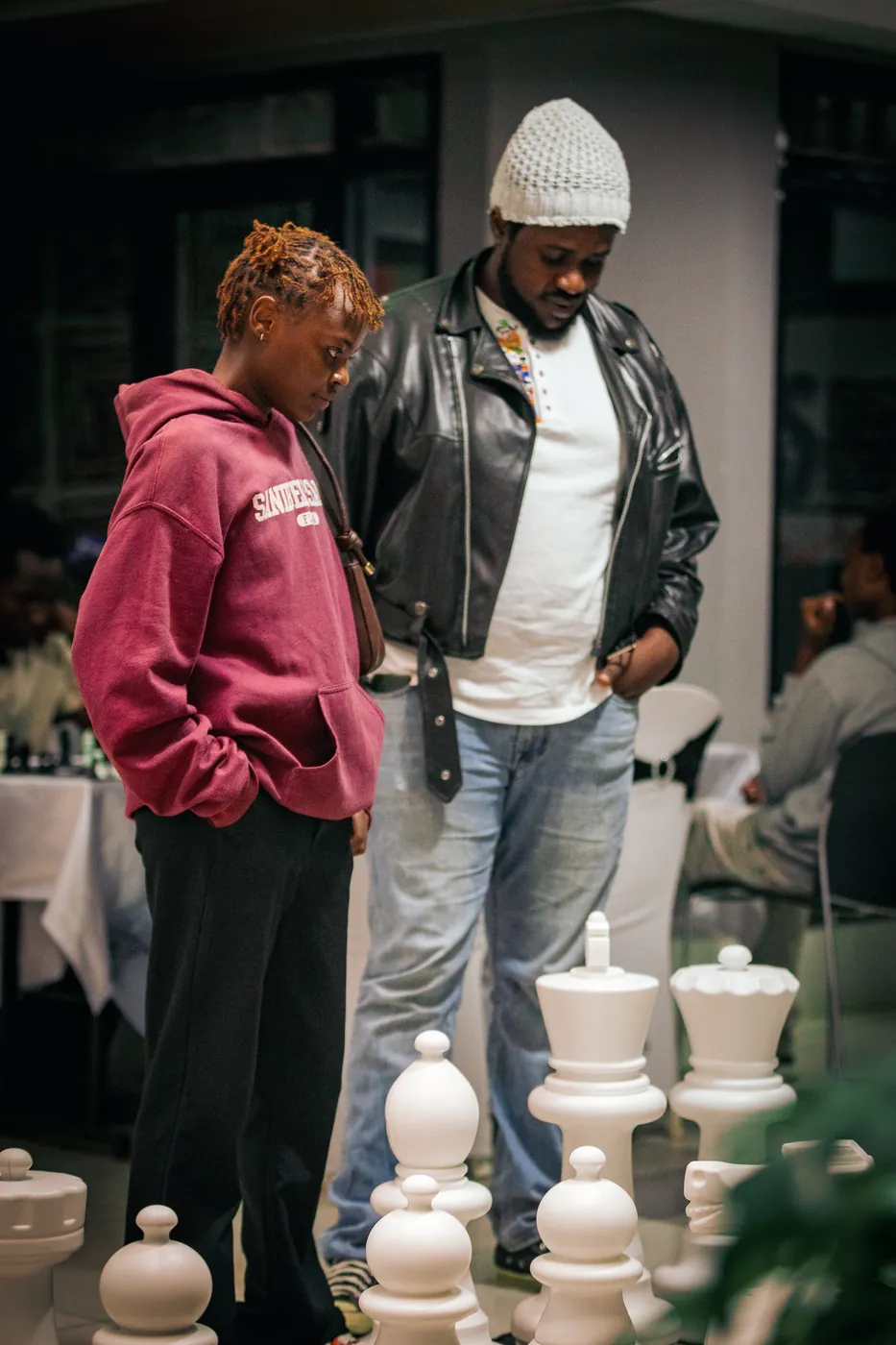A girl in a maroon hoodie and a man look over giant floor chess pieces