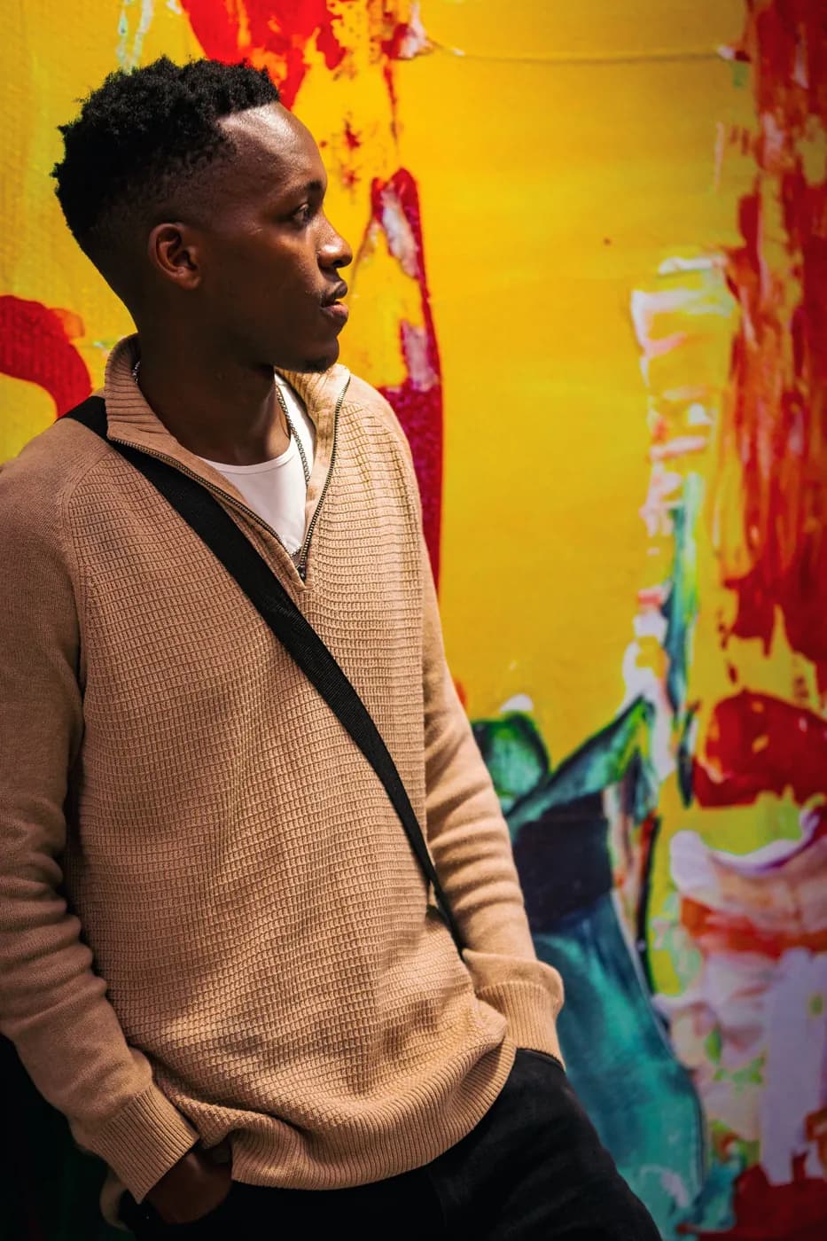 A young man in a beige quarter-zip looks across the room in front of a yellow and red mural