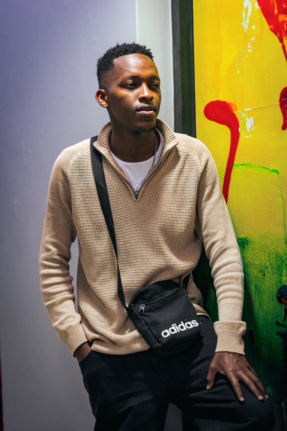 A young man in a beige knit quarter-zip stands against a bright yellow and red painted mural