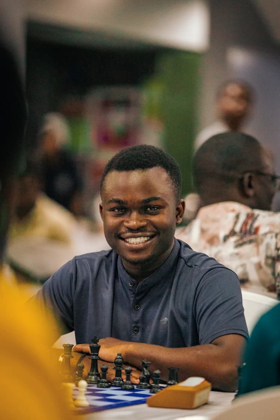A smiling young man in a navy shirt sits behind the black pieces on a blue board