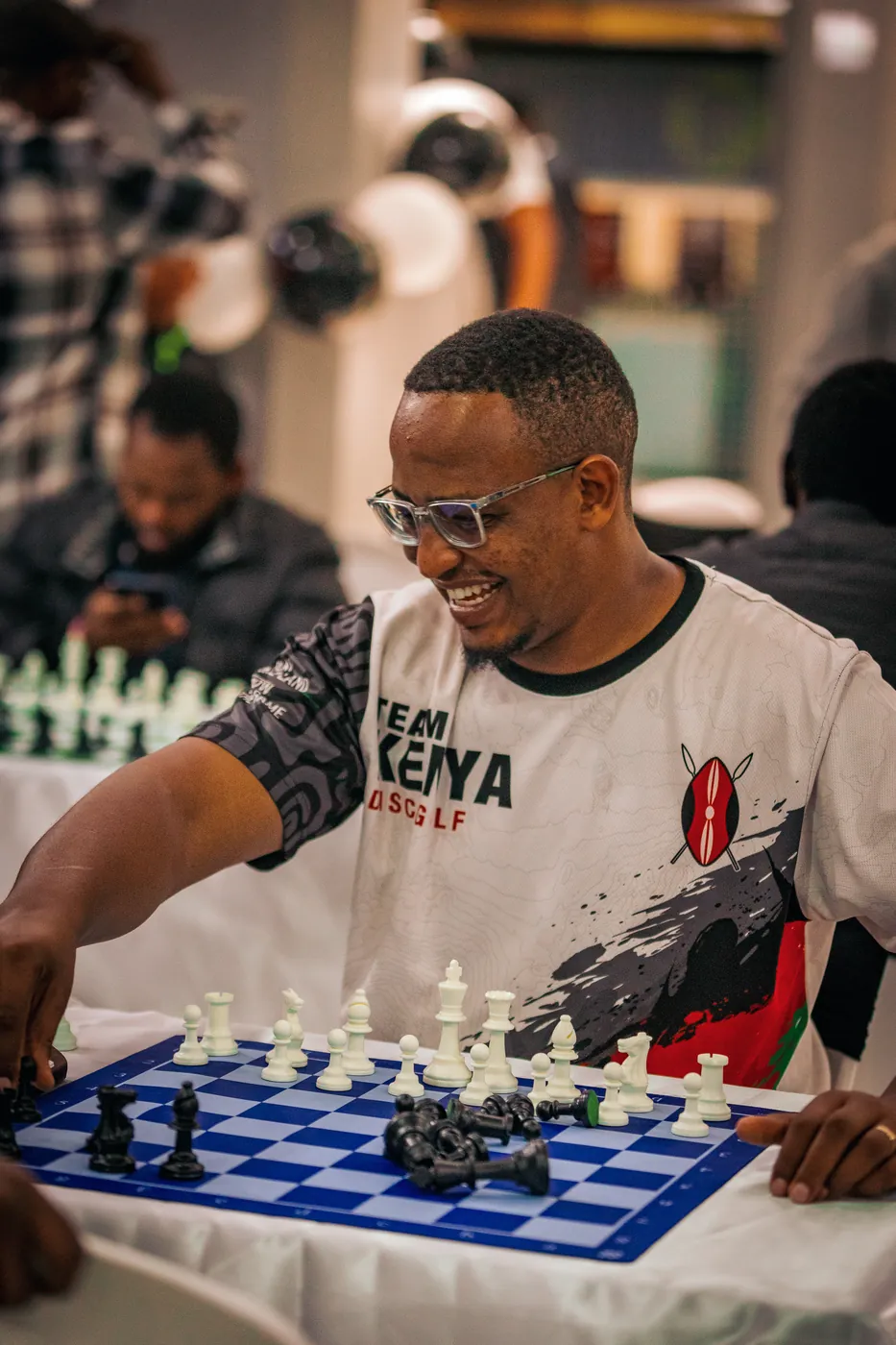 A player in a white Team Kenya tee and clear glasses smiles mid-game