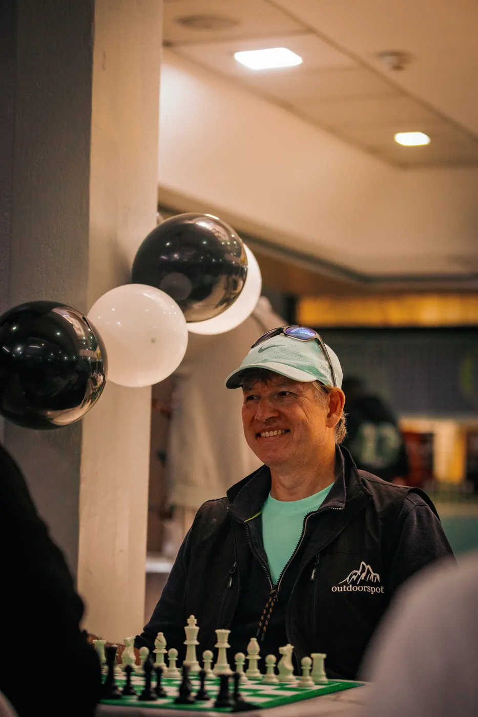 An older player in a white cap and black jacket smiles at the board, black and white balloons behind